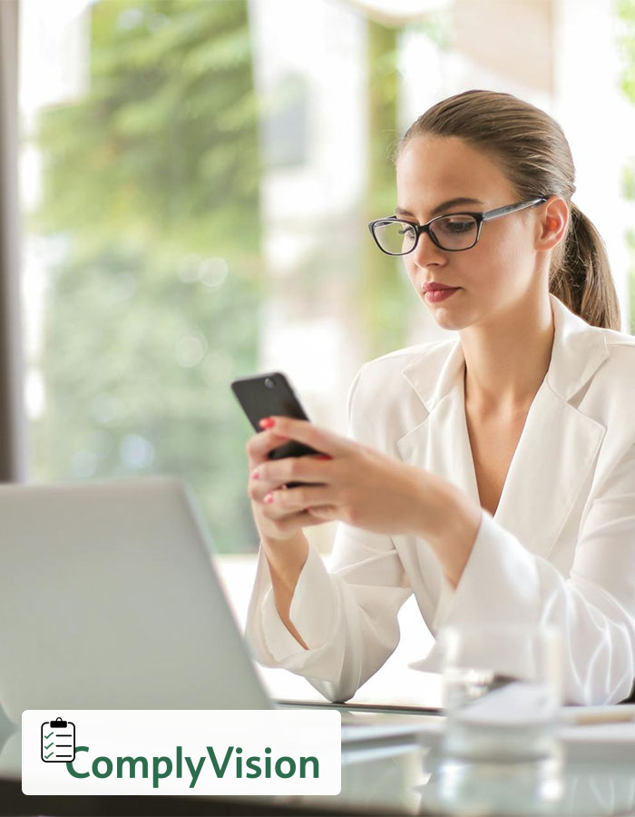 Professional reviewing a smartphone beside a laptop, with the ComplyVision logo in the lower-left corner.